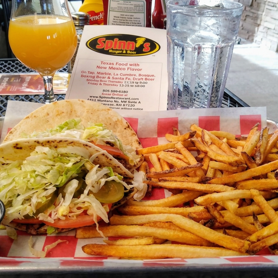 Two loaded tacos with shredded lettuce and tomatoes, fries, dipping sauce, orange drink, and water on an outdoor restaurant table.